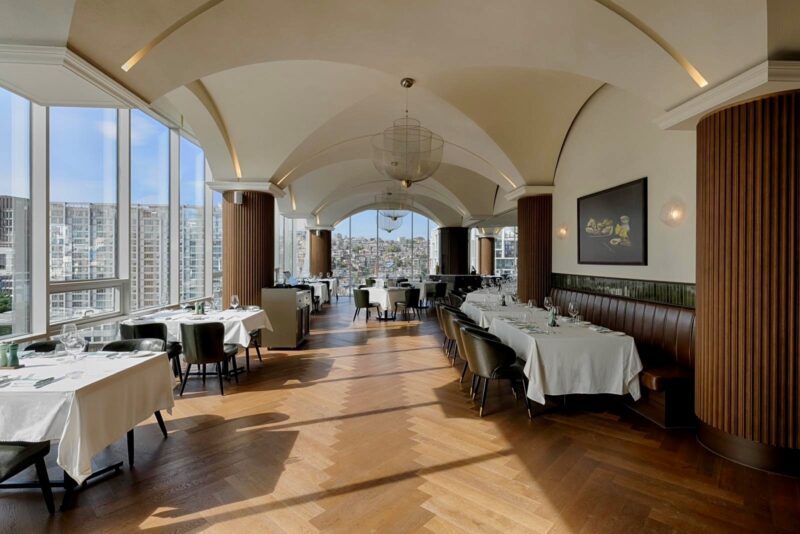 Interior of a restaurant with large windows, arched ceiling, and neatly set tables overlooking a cityscape